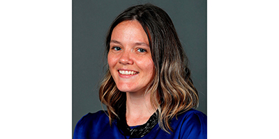 Portrait of a smiling faculty member with shoulder-length brown hair wearing a blue top against a dark background.